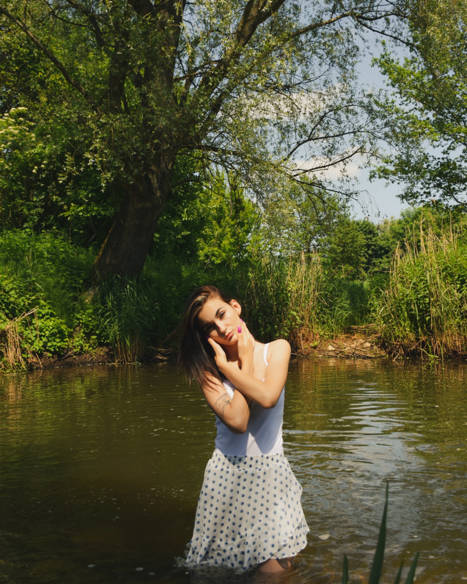 Kobieta w białej sukience w niebieskie kropki stoi w rzece, z rękami skrzyżowanymi pod brodą, w tle zielona roślinność i drzewa.