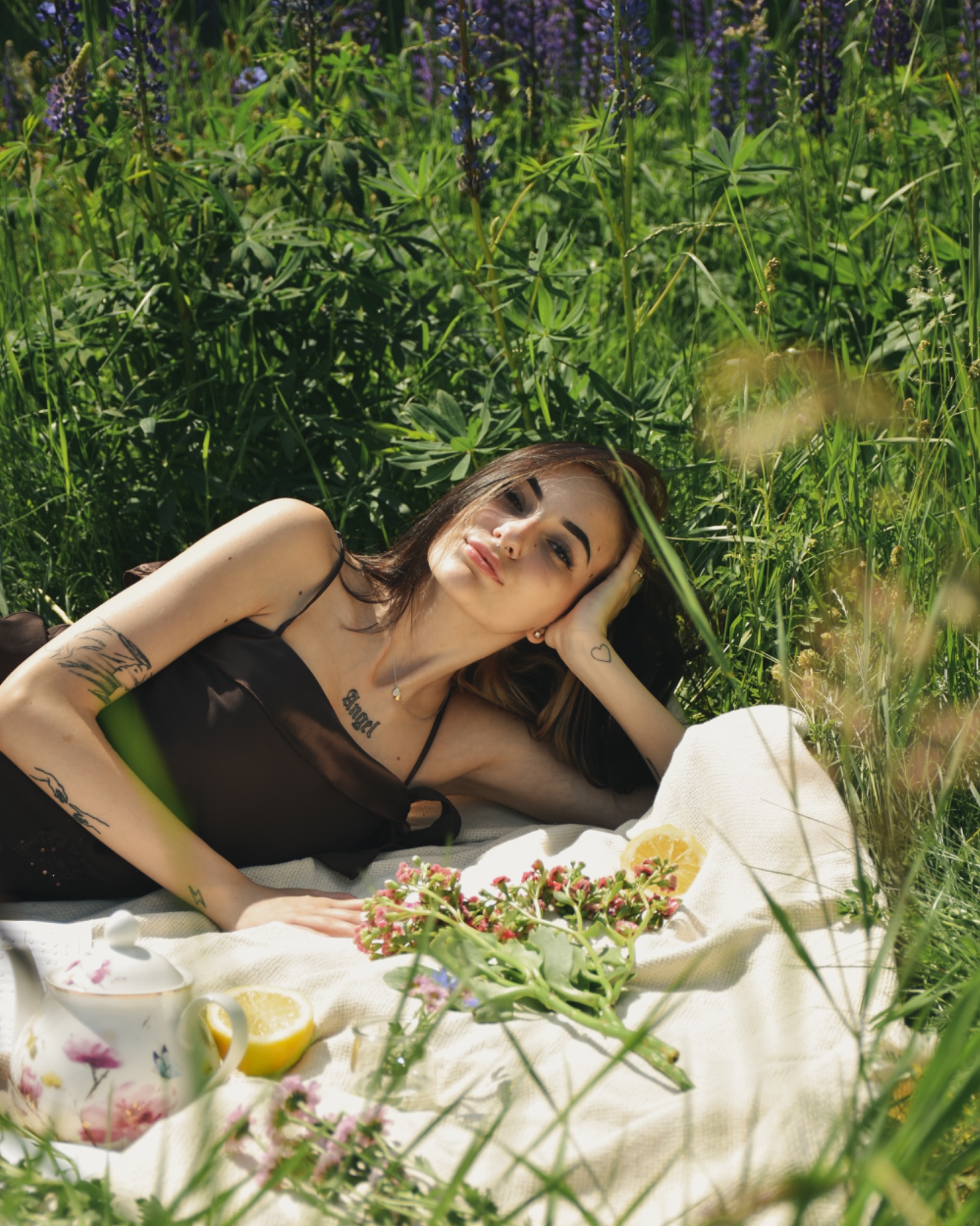 Młoda kobieta w brązowej sukience leży na kocu w wysokiej trawie z kwiatami i cytrynami, obok stoi porcelanowy czajnik w kwiaty.