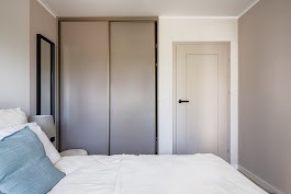 Beżowa szafa przesuwna z lustrem w sypialni, widoczna z perspektywy łóżka. Minimalistyczny design, jasne ściany, czarne detale ramy lustra i klamki.