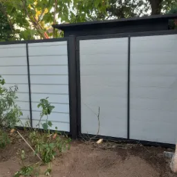 Nowoczesna brama wjazdowa z poziomych paneli w kolorze jasnoszarym, z czarnymi, pionowymi słupkami i ramą, widoczna z zewnątrz posesji, z elementami roślinności w tle i na pierwszym planie.