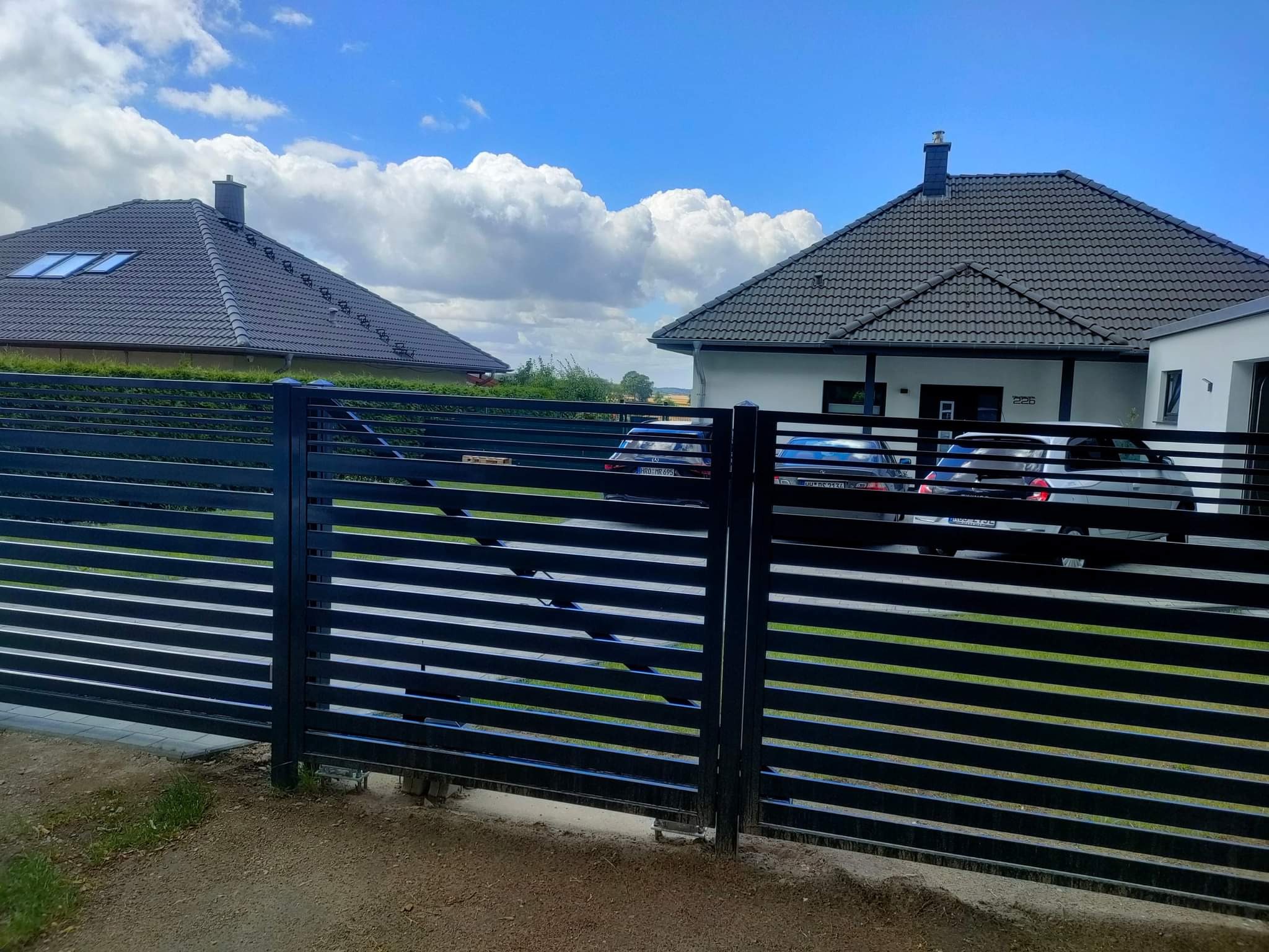 Nowoczesna, pozioma brama przesuwna z czarnych, metalowych profili na tle dwóch domów jednorodzinnych i błękitnego nieba z chmurami.