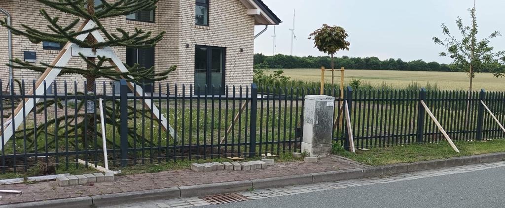 Nowo zamontowane, czarne, metalowe ogrodzenie sztachetowe przed domem z jasnej cegły. Widoczne podpory drewniane stabilizujące konstrukcję, ułożone kostki brukowe i drzewo z dodatkowym wsparciem.