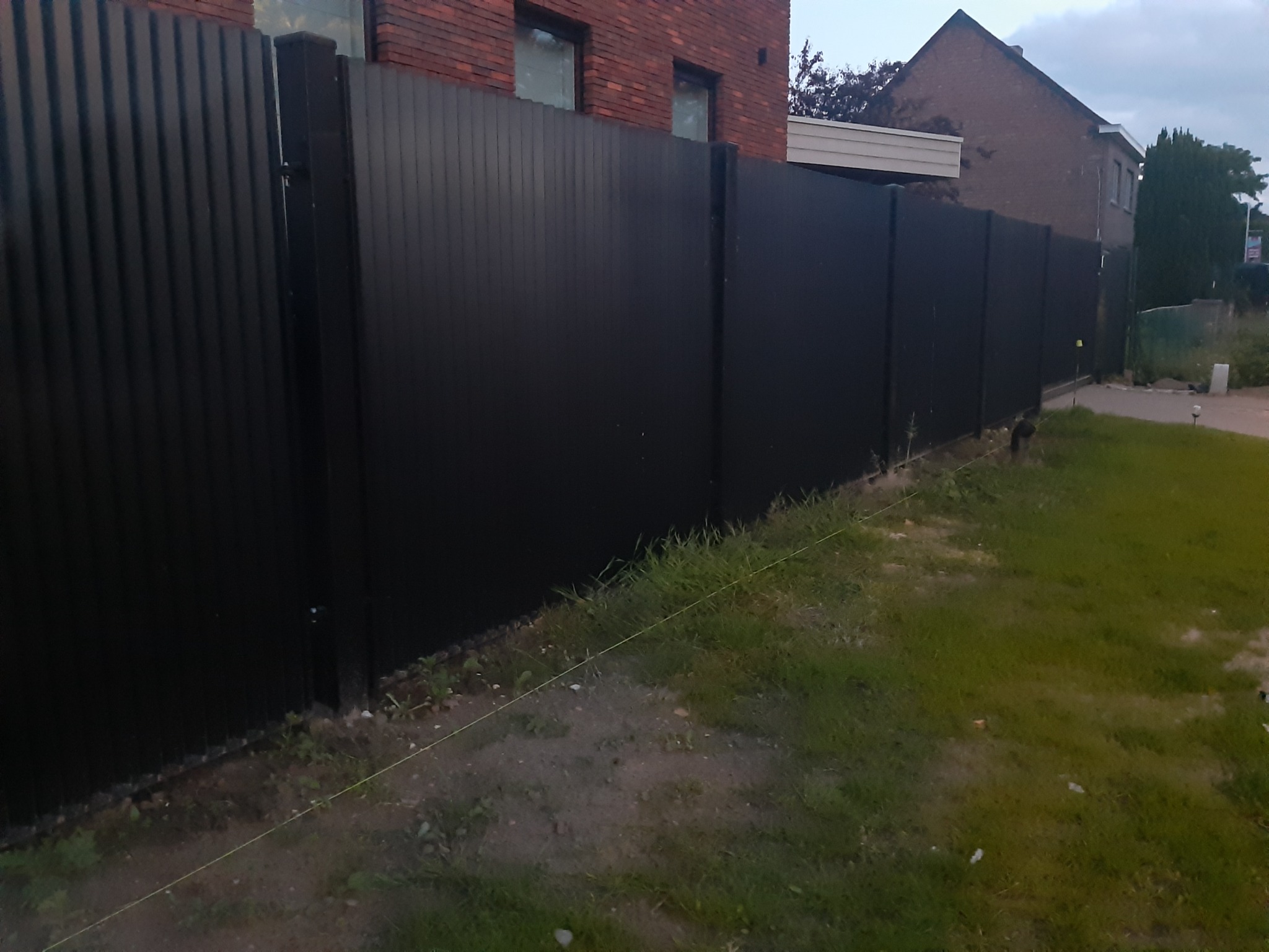 Długi, czarny, metalowy płot panelowy z widocznymi słupkami, rozciągnięty wzdłuż trawnika przed domem z czerwonej cegły.