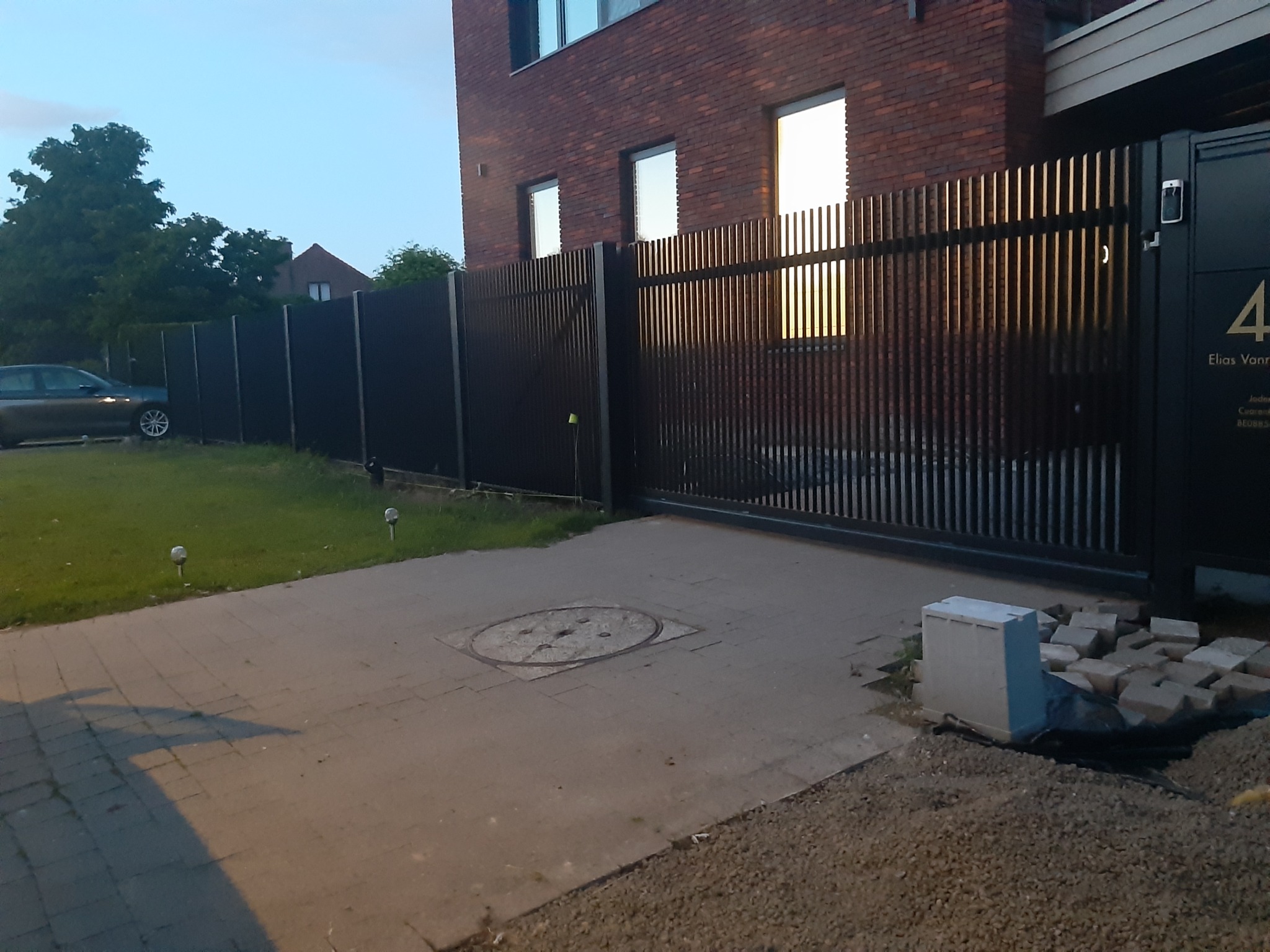 Nowoczesne, pionowe, metalowe ogrodzenie z przesuwną bramą wjazdową, widoczne niedokończone prace brukarskie i sterta kostki brukowej.