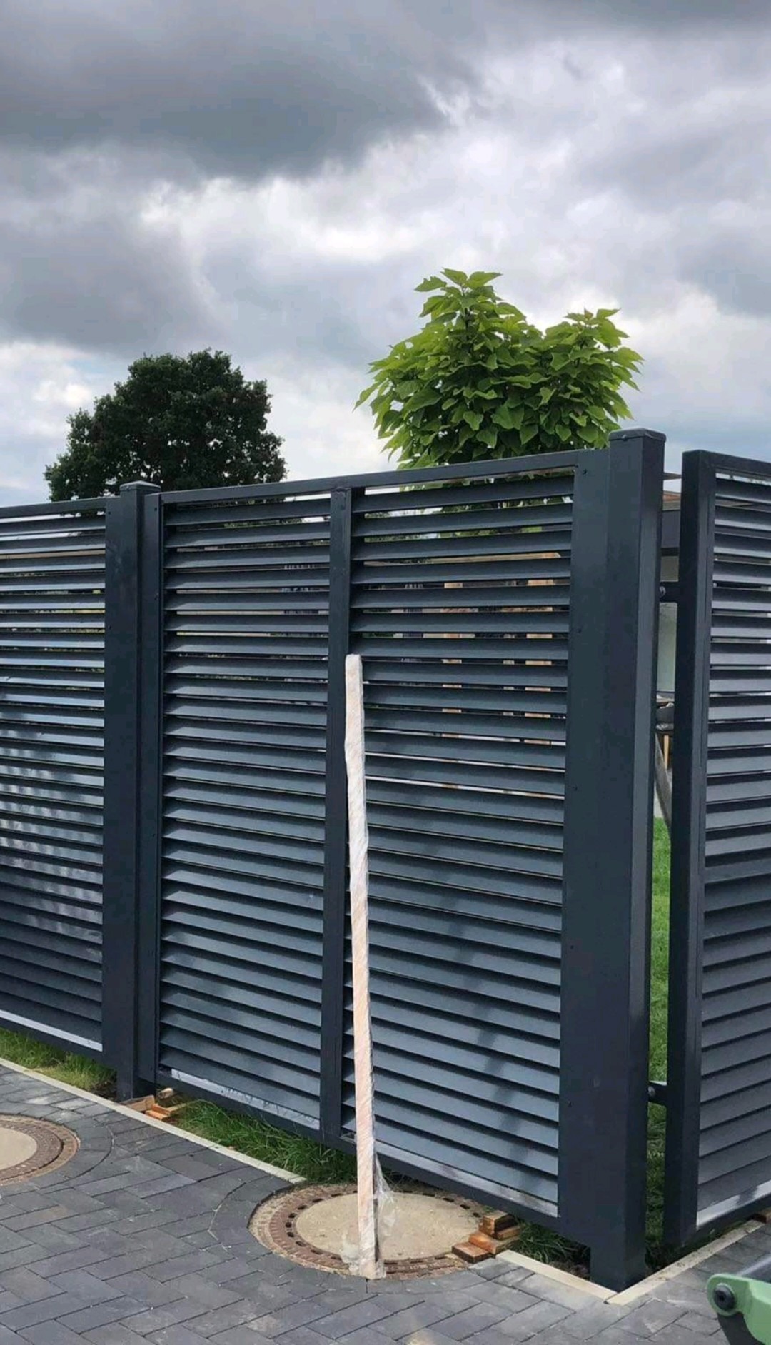 Nowoczesne, szare ogrodzenie panelowe z poziomymi lamelami w Kamieniu Małym, widoczne elementy montażowe w folii ochronnej, kostka brukowa i studzienki kanalizacyjne w tle.