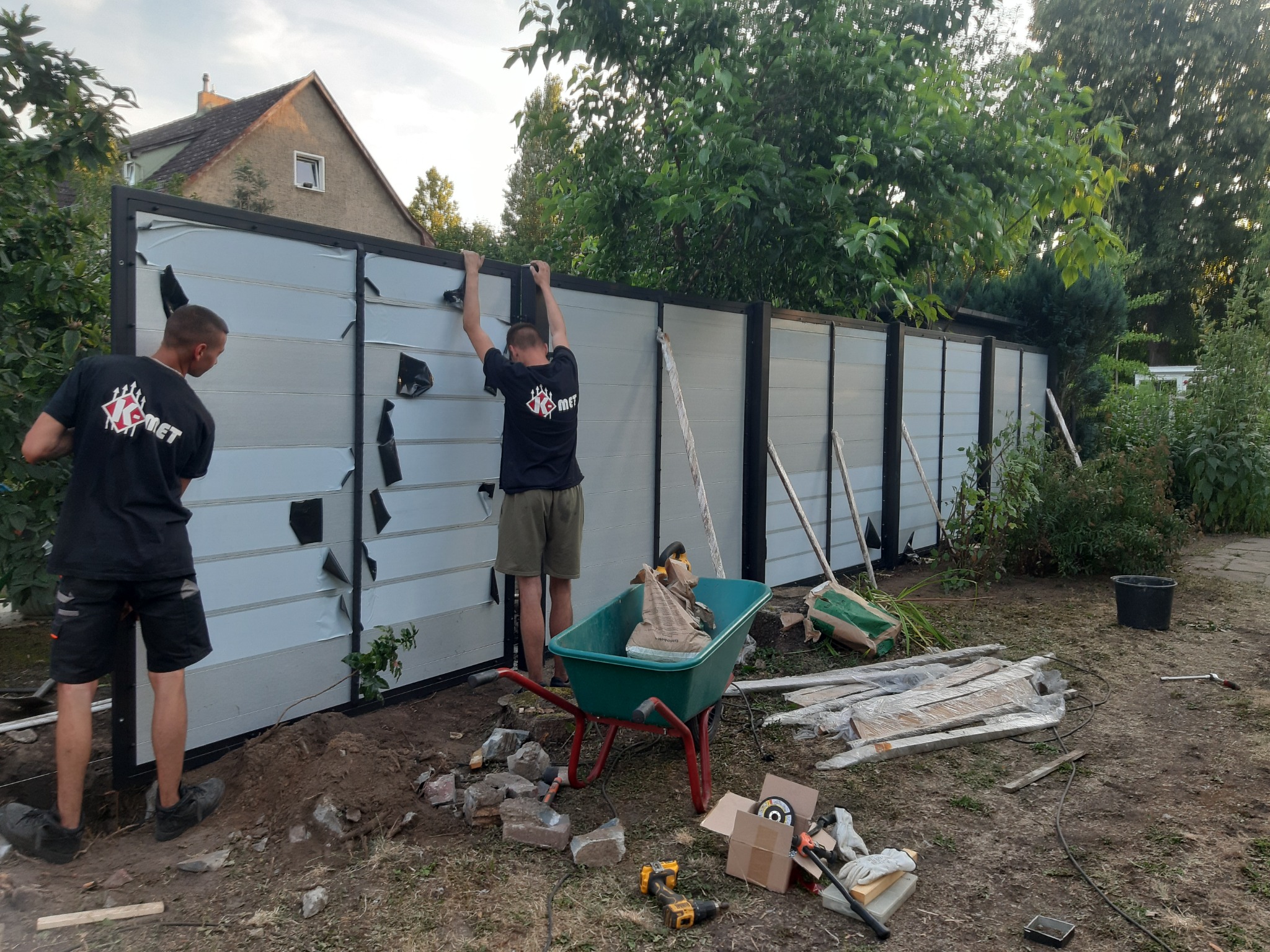 Dwóch mężczyzn w trakcie montażu ogrodzenia z paneli z czarną ramą, widoczne narzędzia, taczka z cementem, oraz częściowo zdjęta folia ochronna z paneli. W tle dom i zieleń.