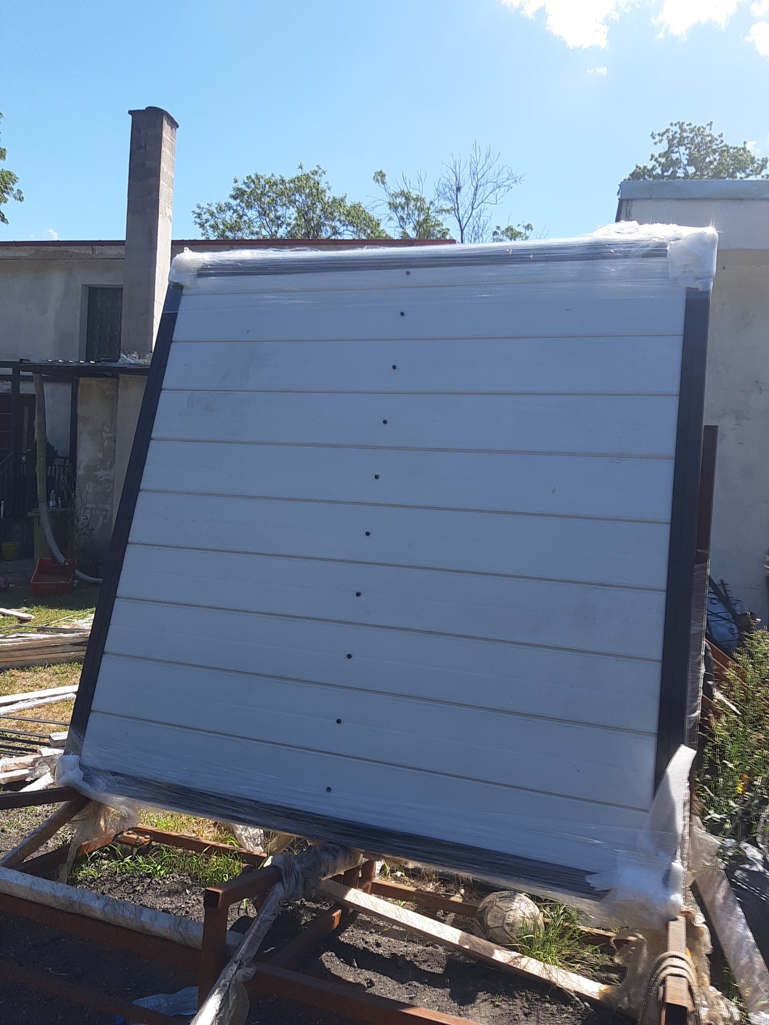 Element ogrodzenia panelowego, zabezpieczony folią, oparty o metalową konstrukcję na trawniku z widoczną piłką do gry, na tle budynku z kominem.