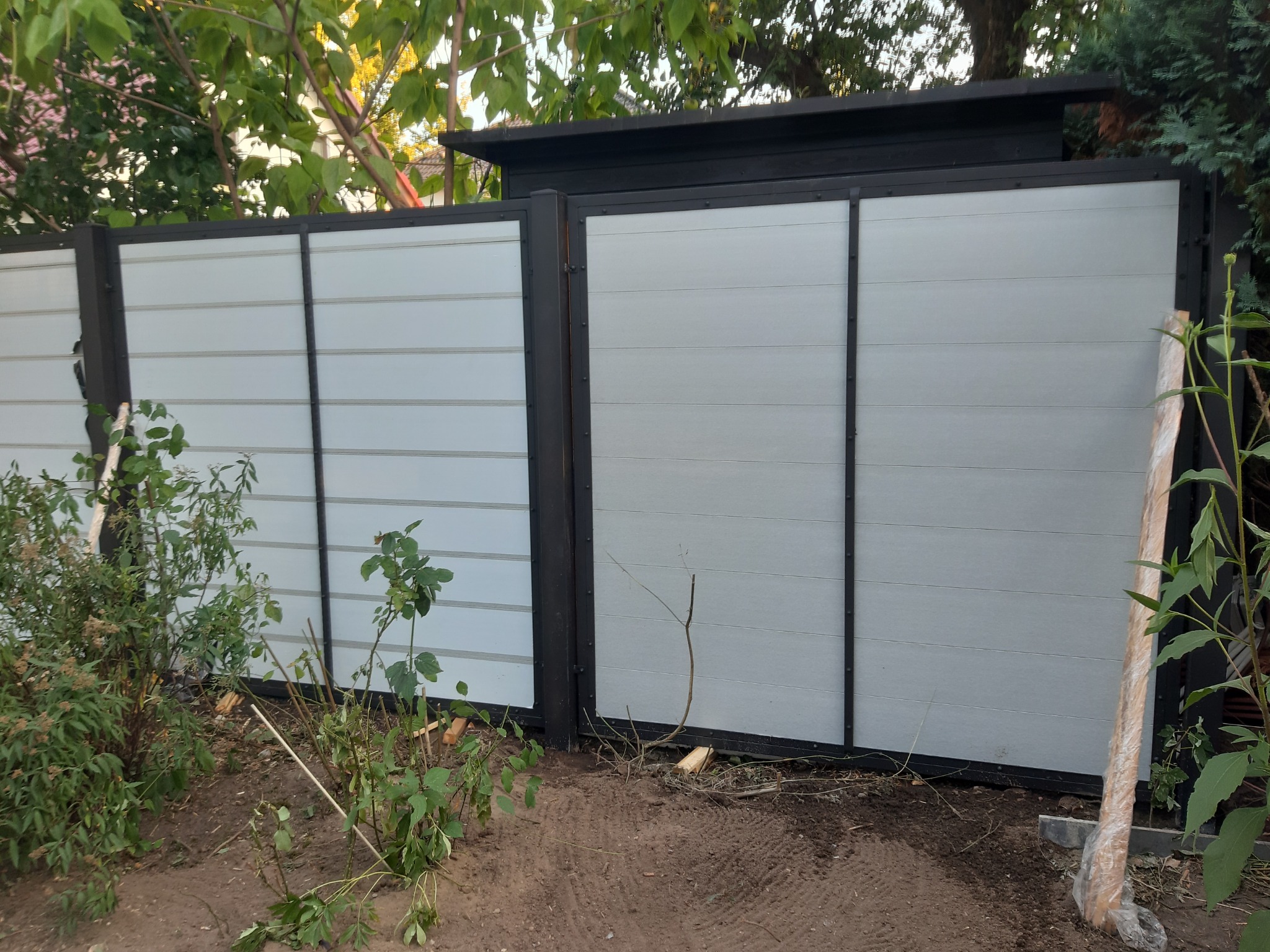 Nowoczesna brama wjazdowa z poziomych paneli w kolorze jasnoszarym, z czarnymi, pionowymi słupkami i ramą, widoczna z zewnątrz posesji, z elementami roślinności w tle i na pierwszym planie.