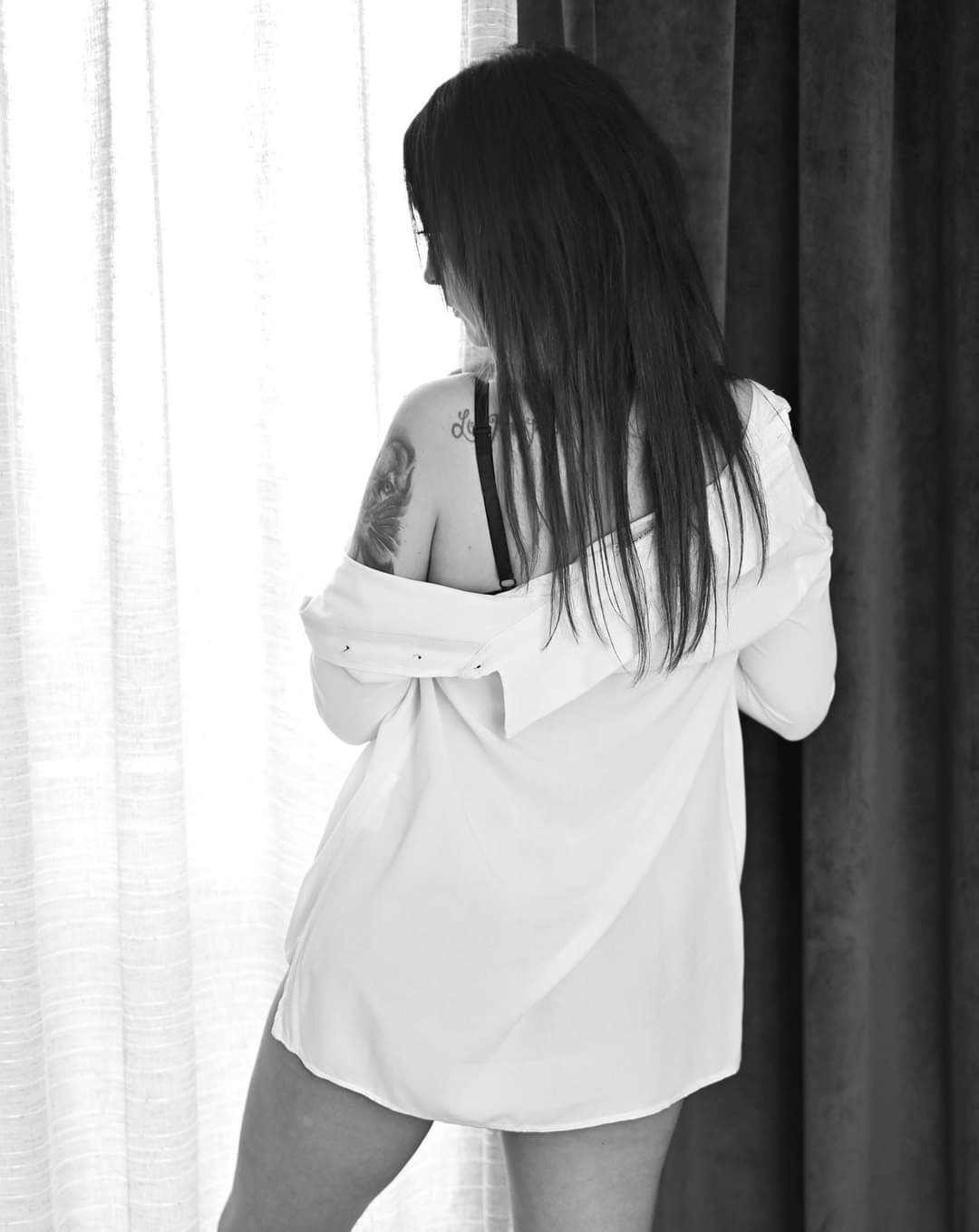 Czarno-białe zdjęcie kobiety z odsłoniętym ramieniem, widok z tyłu, w rozpiętej białej koszuli, stojącej przy oknie z firanką i zasłoną, widoczny tatuaż na ramieniu.