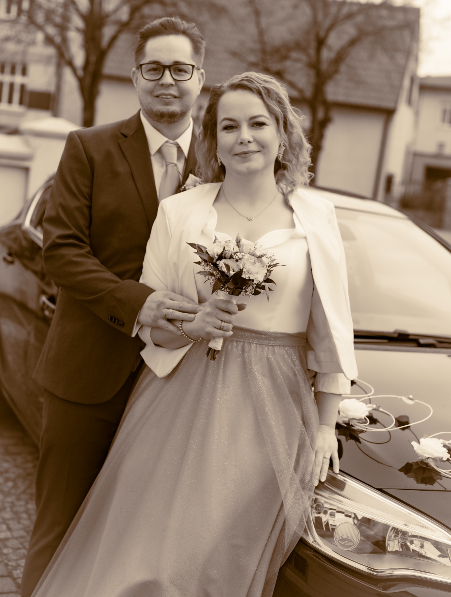 Para młoda w strojach ślubnych pozuje przy udekorowanym samochodzie, panna młoda trzyma bukiet, zdjęcie w sepii.