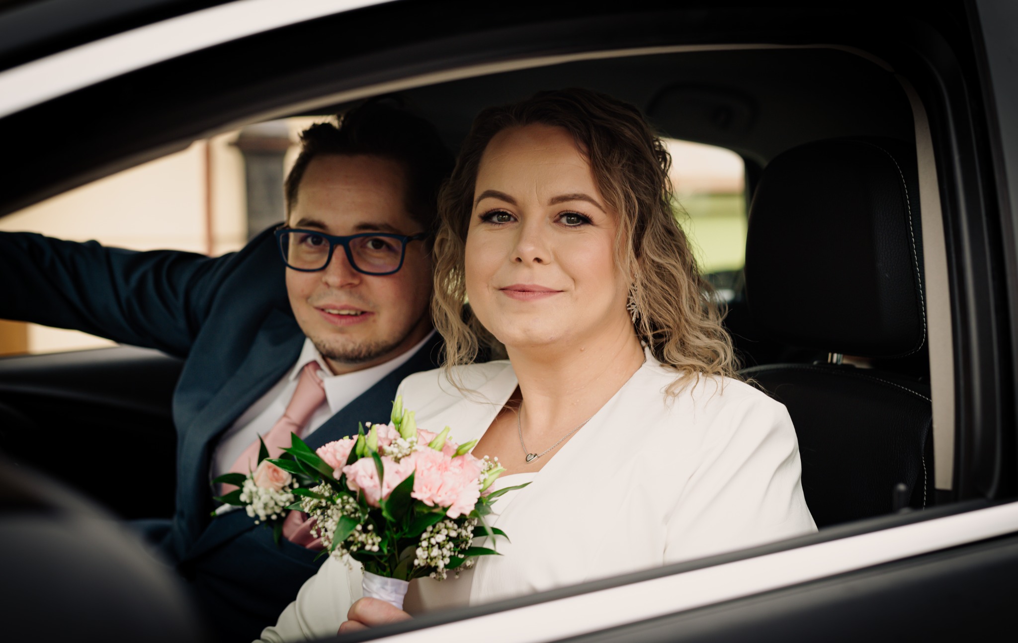 Para młoda siedząca w samochodzie: panna młoda trzyma bukiet różowych kwiatów, pan młody w garniturze z różowym krawatem, oboje uśmiechnięci.
