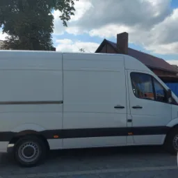 Biały samochód dostawczy typu furgon marki Mercedes z przesuwanymi drzwiami bocznymi, zaparkowany na tle budynku z ceglanym kominem i błękitnego ogrodzenia.