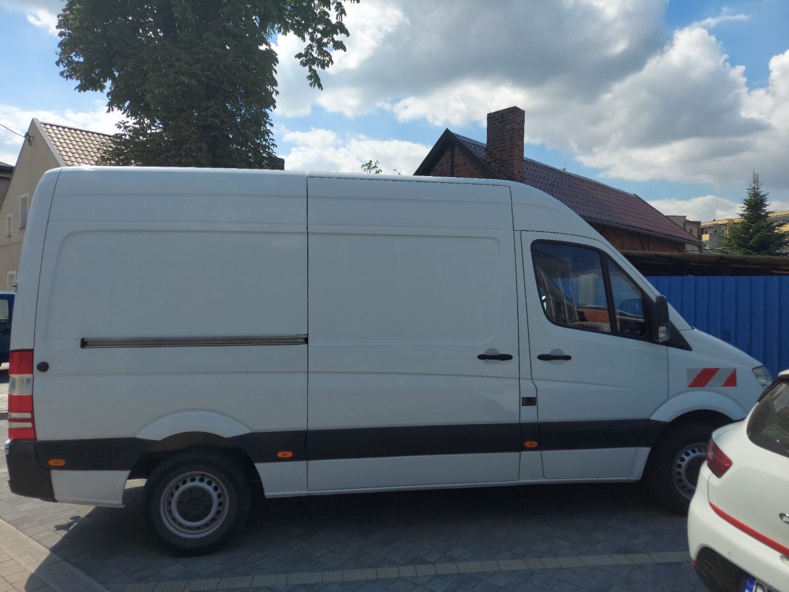 Biały samochód dostawczy typu furgon marki Mercedes z przesuwanymi drzwiami bocznymi, zaparkowany na tle budynku z ceglanym kominem i błękitnego ogrodzenia.