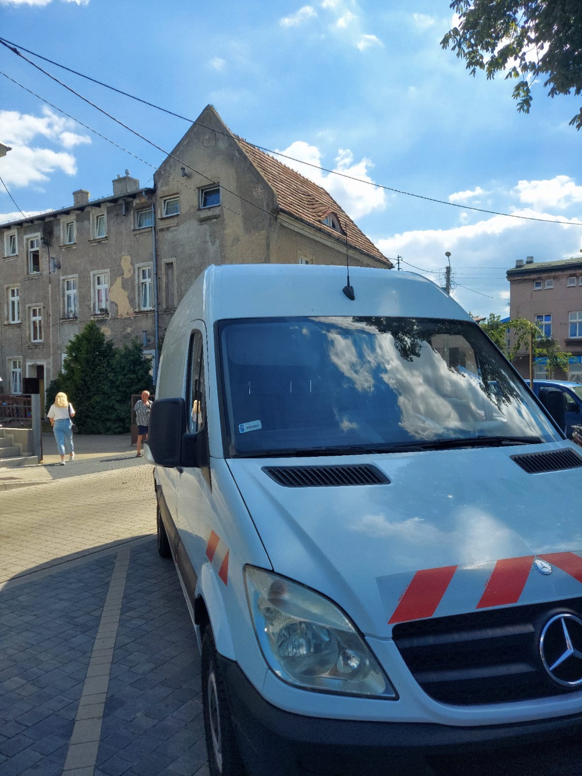 Biały bus Mercedes z pomarańczowymi pasami odblaskowymi na tle miejskiej zabudowy i błękitnego nieba z chmurami odbijającymi się w przedniej szybie.