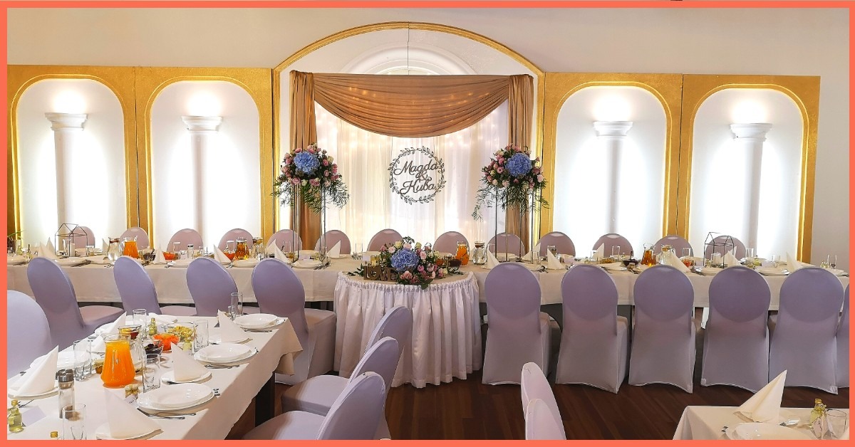 Uroczysta sala bankietowa z dekoracją kwiatową, personalizowanym napisem Magda & Kuba, stołami nakrytymi białymi obrusami i krzesłami w pokrowcach w kolorze liliowym.