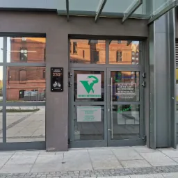 Wejście do przychodni dla zwierząt z logo zielonego węża w kielichu, tabliczką z adresem i godzinami otwarcia, widok z zewnątrz, w tle budynki odbijające się w szybach.