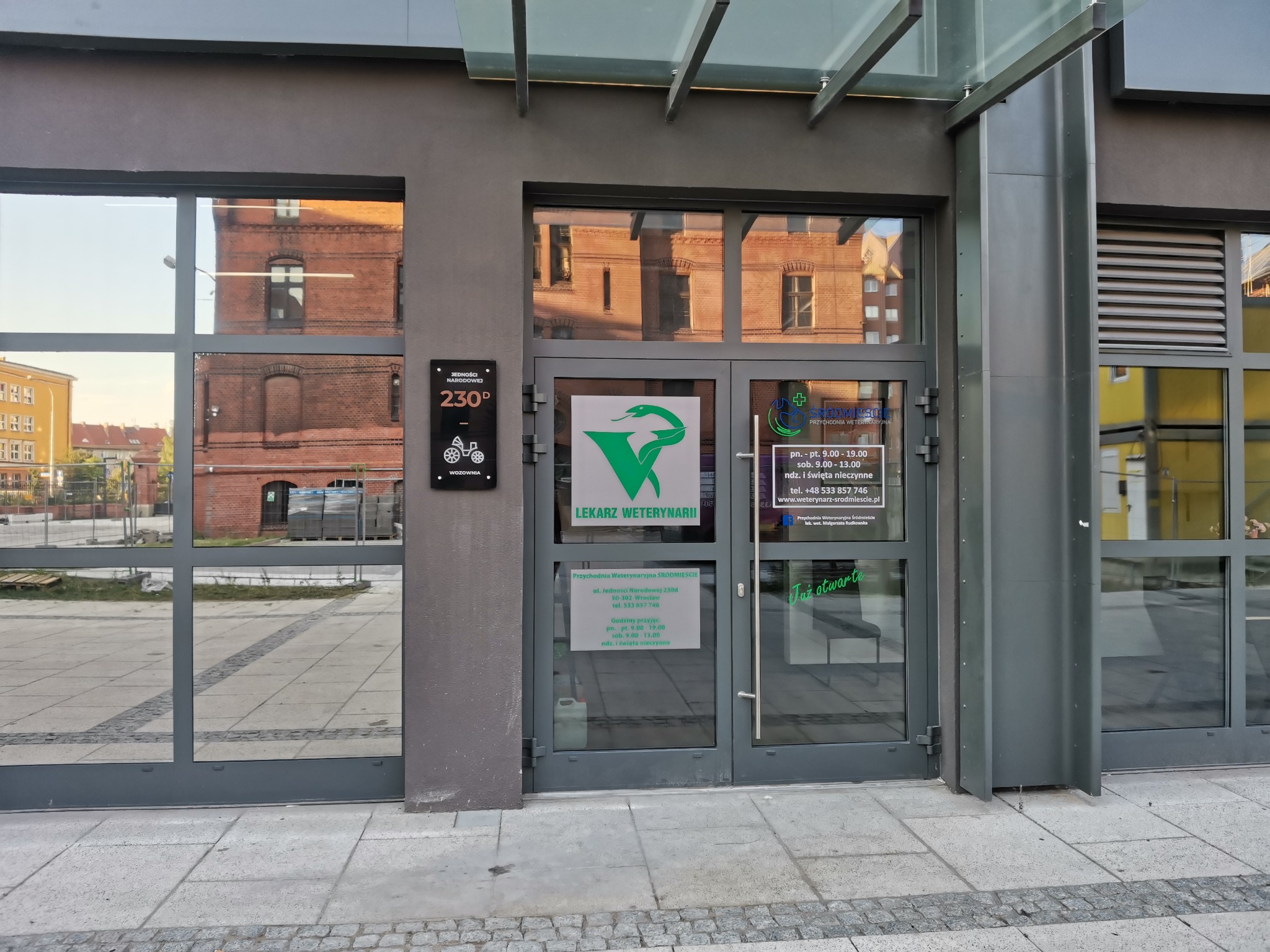 Wejście do przychodni dla zwierząt z logo zielonego węża w kielichu, tabliczką z adresem i godzinami otwarcia, widok z zewnątrz, w tle budynki odbijające się w szybach.