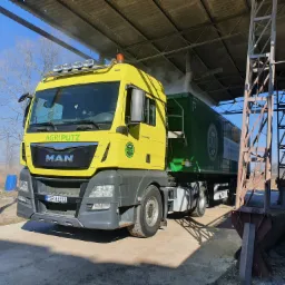 Żółta ciężarówka AgriPutz pod zadaszeniem podczas rozładunku, z widocznym pyłem unoszącym się z naczepy. W tle drzewa bez liści i błękitne niebo.