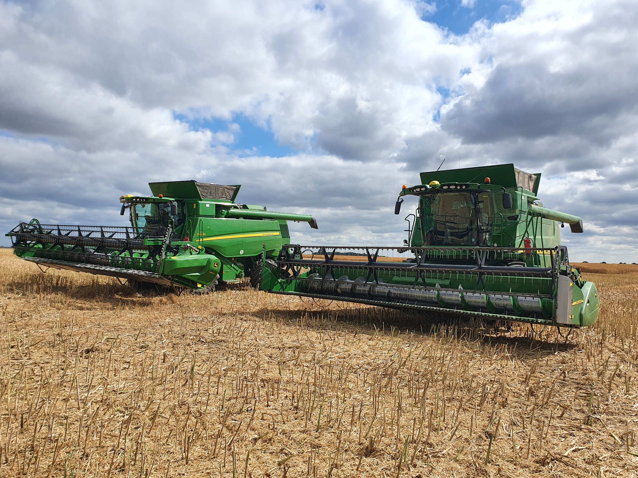 Dwa zielone kombajny zbożowe John Deere na polu po żniwach, pod pochmurnym niebem.