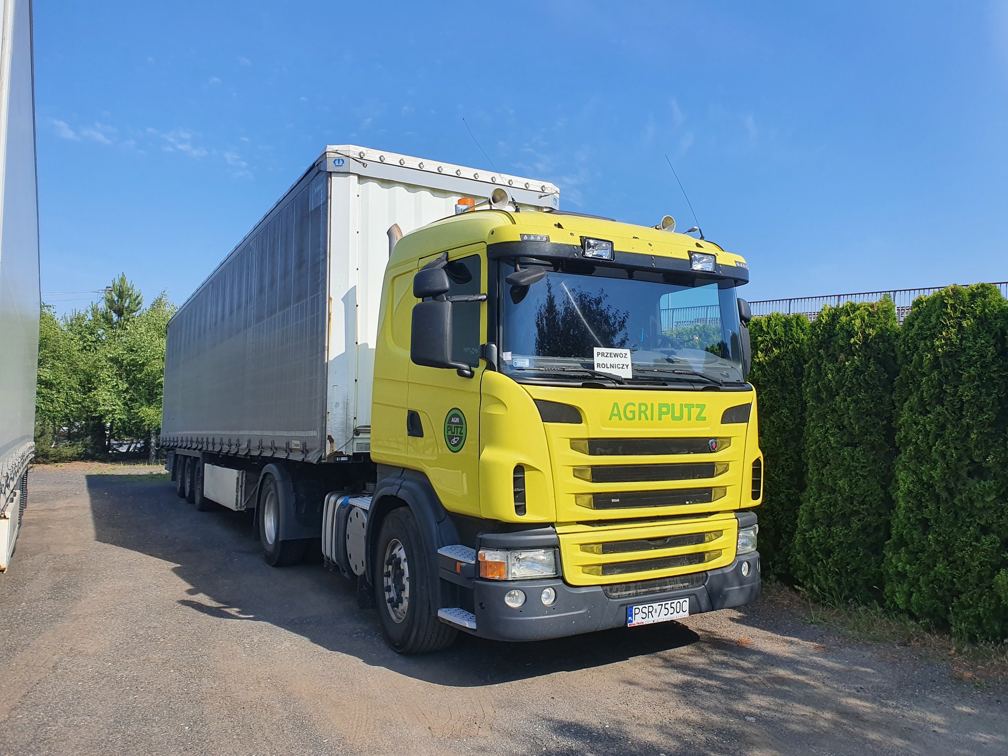 Żółta ciężarówka z naczepą, z napisem AGRI PUTZ, zaparkowana na utwardzonym placu, z zielonym żywopłotem w tle i błękitnym niebem.