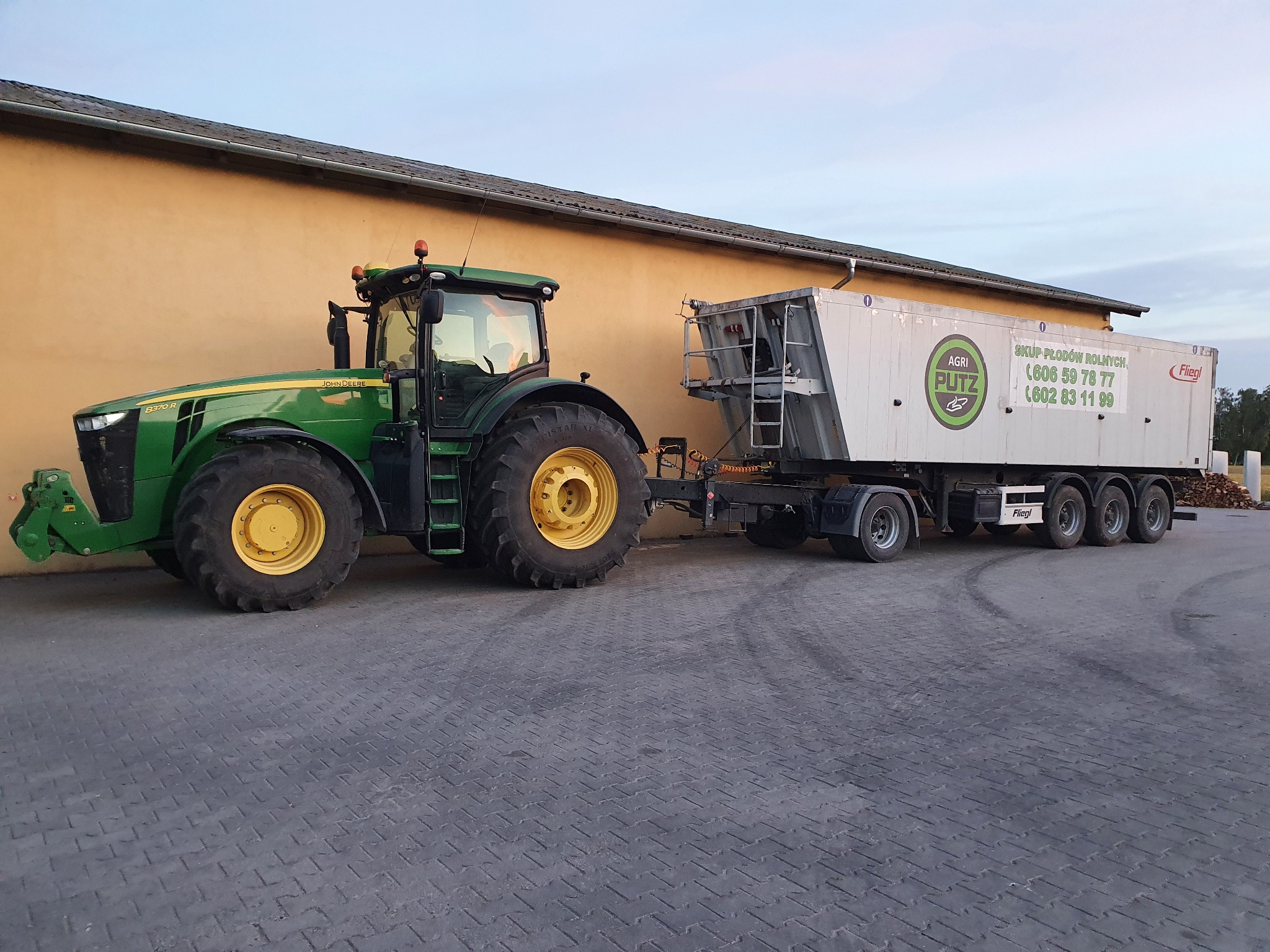 Zielony ciągnik John Deere 8370R z przyczepą do transportu płodów rolnych, zaparkowany na brukowanym placu przed żółtym budynkiem, z widocznym logo i numerami telefonów firmy skupującej płody rolne...