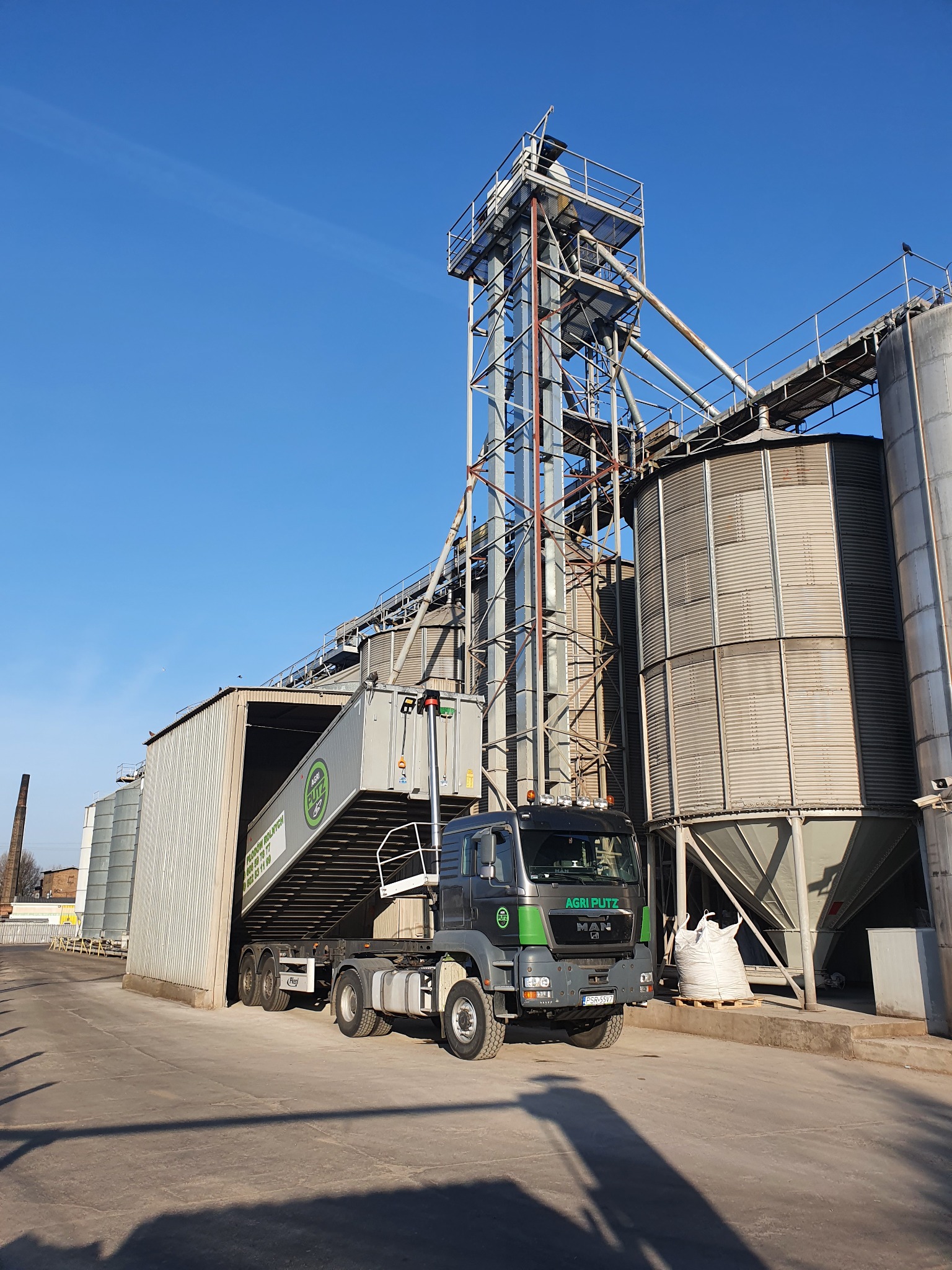 Ciężarówka Agri Putz z naczepą pod silosami zbożowymi w słoneczny dzień.
