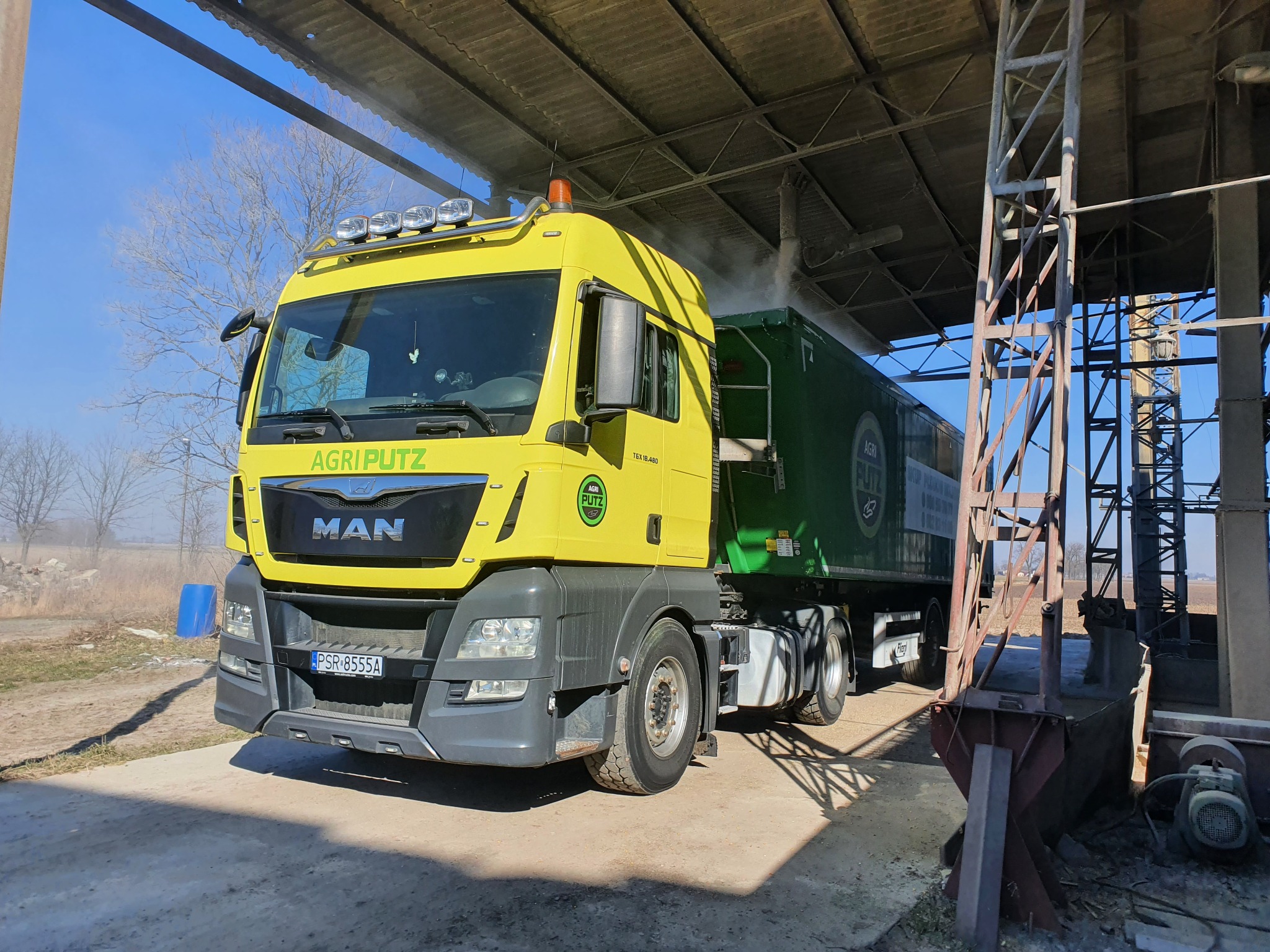 Żółta ciężarówka AgriPutz pod zadaszeniem podczas rozładunku, z widocznym pyłem unoszącym się z naczepy. W tle drzewa bez liści i błękitne niebo.