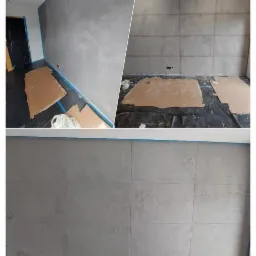 Ściana w szarym odcieniu z efektem betonu architektonicznego, podzielona na kwadratowe panele, zabezpieczona niebieską taśmą malarską przy suficie, podłoga zabezpieczona folią i kartonami.