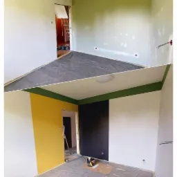 Dwa ujęcia tego samego pokoju: górne przed malowaniem i szpachlowaniem, z widocznymi przewodami i zakrytą podłogą; dolne po malowaniu, z żółtą i czarną ścianą, zielonym pasem pod sufitem, zakrytą...