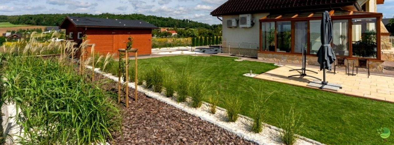 Widok na zadbany ogród z równo przystrzyżonym trawnikiem, ozdobnymi trawami wzdłuż rabat wysypanych korą i białym grysem, drewnianą altaną w tle oraz domem z przeszkloną werandą i klimatyzacją...