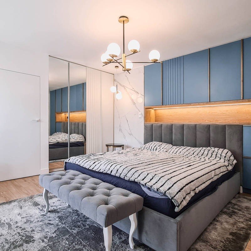Nowoczesna sypialnia z szarą tapicerowaną ławką przed łóżkiem, niebieskimi panelami ściennymi i kulistym żyrandolem w stylu mid-century.