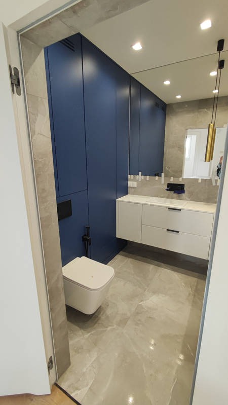 Nowoczesna łazienka z szafkami w odcieniach granatu i bieli, marmurowa podłoga, oświetlenie punktowe i minimalistyczna armatura, widok zza futryny.