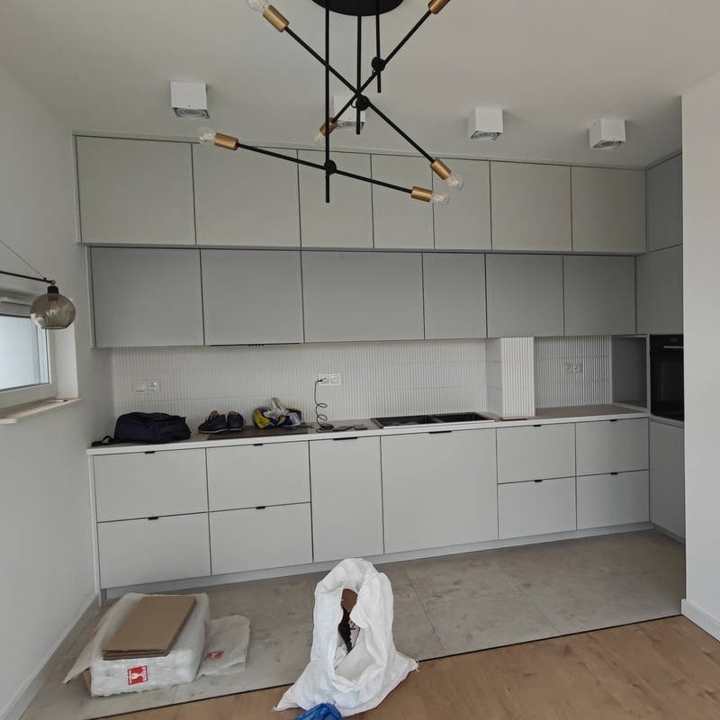Jasna, minimalistyczna kuchnia z szarymi szafkami i czarnym, nowoczesnym oświetleniem. Widoczne detale wykończenia i pozostałości po montażu.