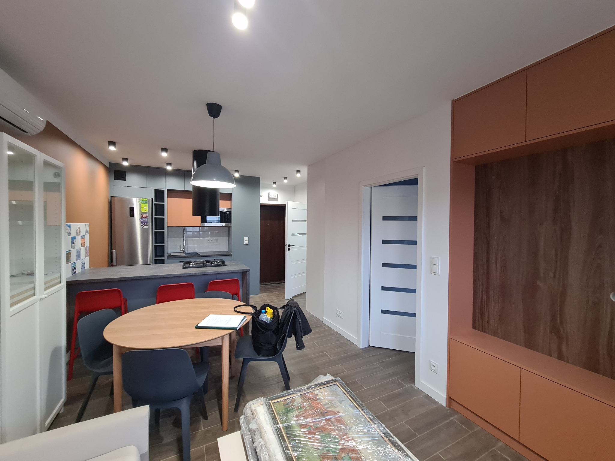 Nowoczesna aranżacja otwartej przestrzeni: jadalnia z okrągłym stołem, kuchnia z szarym blatem i zabudową w odcieniach brązu, białe drzwi z poziomymi frezami.