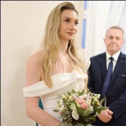 Olga Umyszkiewicz - Panna młoda w białej sukni z bukietem kwiatów, obok mężczyzna w garniturze. Ujęcie z uroczystości ślubnej, z delikatnym oświetleniem.