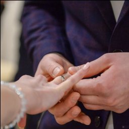 Olga Umyszkiewicz - Zbliżenie dłoni zakładających obrączkę na palec podczas ceremonii ślubnej. Delikatne oświetlenie podkreśla detale biżuterii i fakturę materiału garnituru.