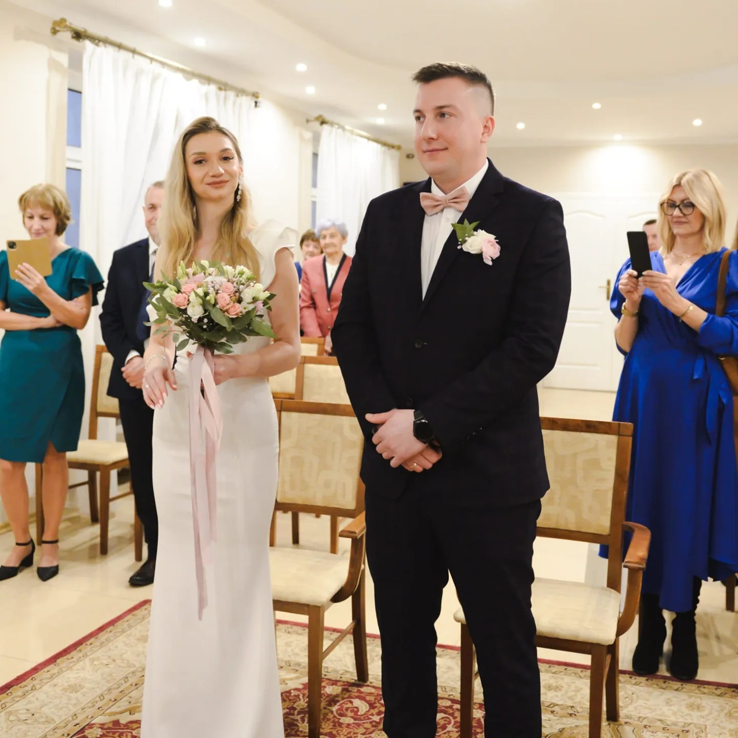 Elegancka para młoda podczas ceremonii ślubnej, panna młoda z bukietem, pan młody w ciemnym garniturze i różowej muszce, goście w tle.