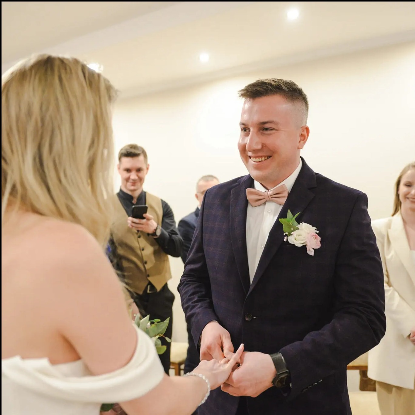 Uśmiechnięty pan młody w bordowym garniturze i muszce, trzymający dłoń panny młodej podczas ceremonii ślubnej w jasnym wnętrzu, z gośćmi w tle.