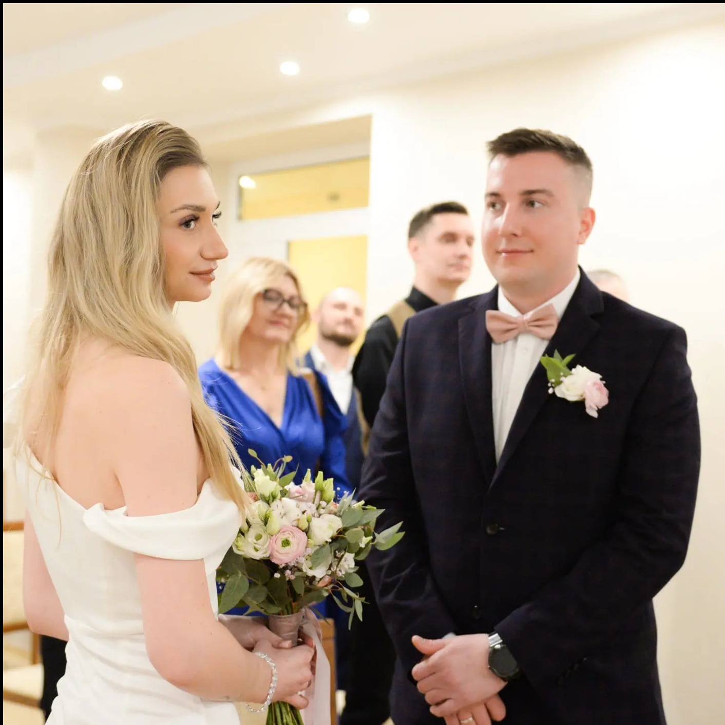 Para młoda podczas ceremonii ślubnej. Panna młoda z bukietem, pan młody w garniturze z muszką. Goście w tle. Jasne wnętrze.