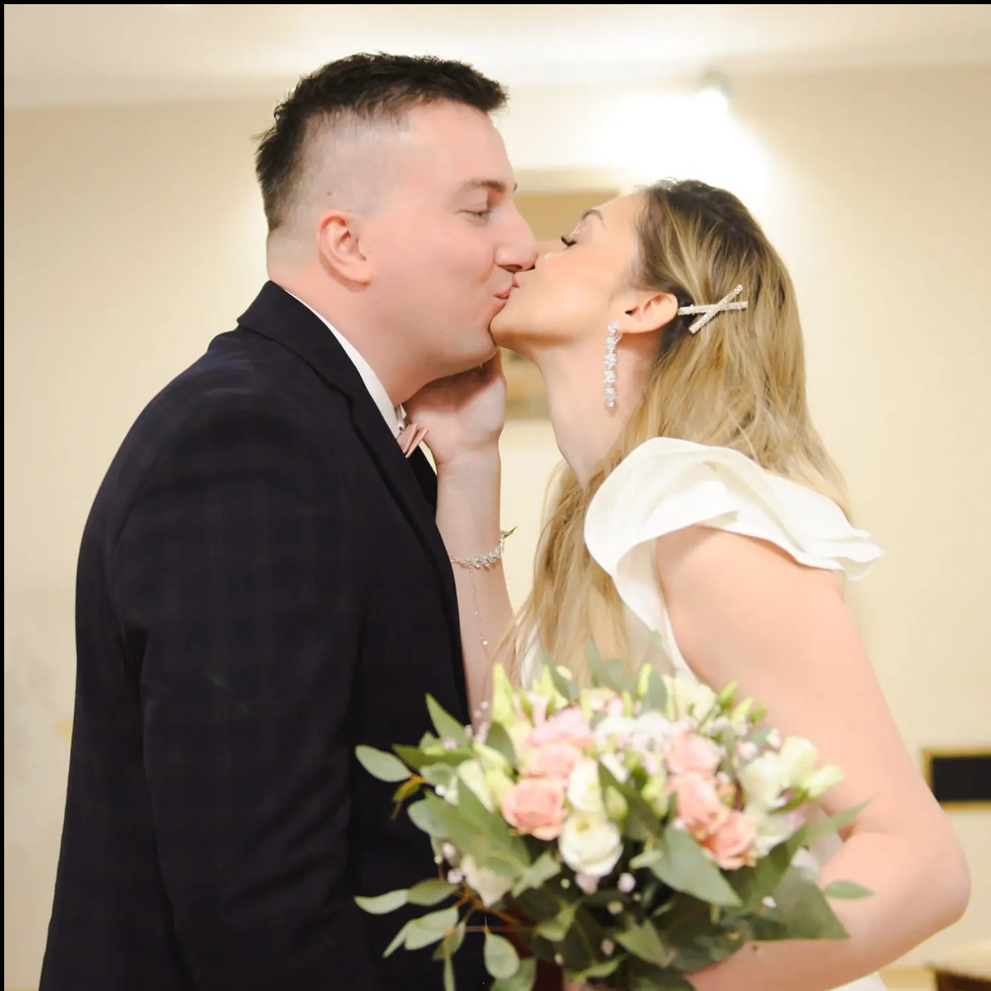 Pocałunek młodej pary podczas ceremonii ślubnej. Panna młoda w białej sukni trzyma bukiet kwiatów. Pan młody w ciemnym garniturze. Delikatne oświetlenie.