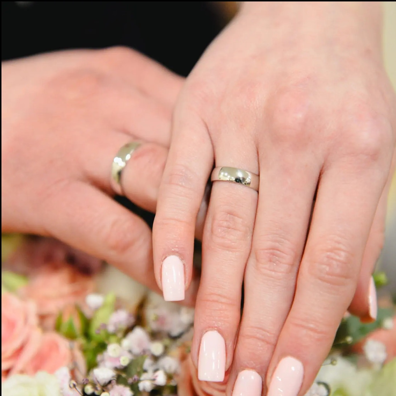 Dłonie pary młodej z obrączkami na tle bukietu ślubnego w delikatnych, pastelowych kolorach. Obrączki srebrne, gładkie. Manicure w odcieniu pudrowego różu.