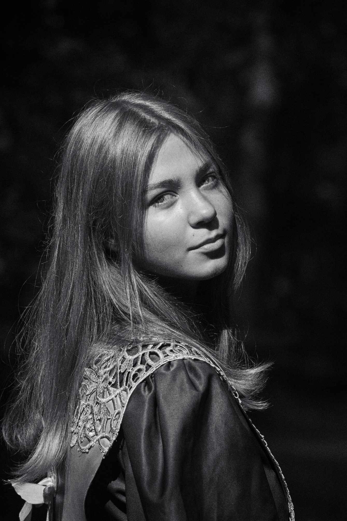 Czarno-białe zdjęcie portretowe młodej kobiety z długimi włosami, spoglądającej przez ramię, z dekoracyjnym wykończeniem sukienki na ramieniu.
