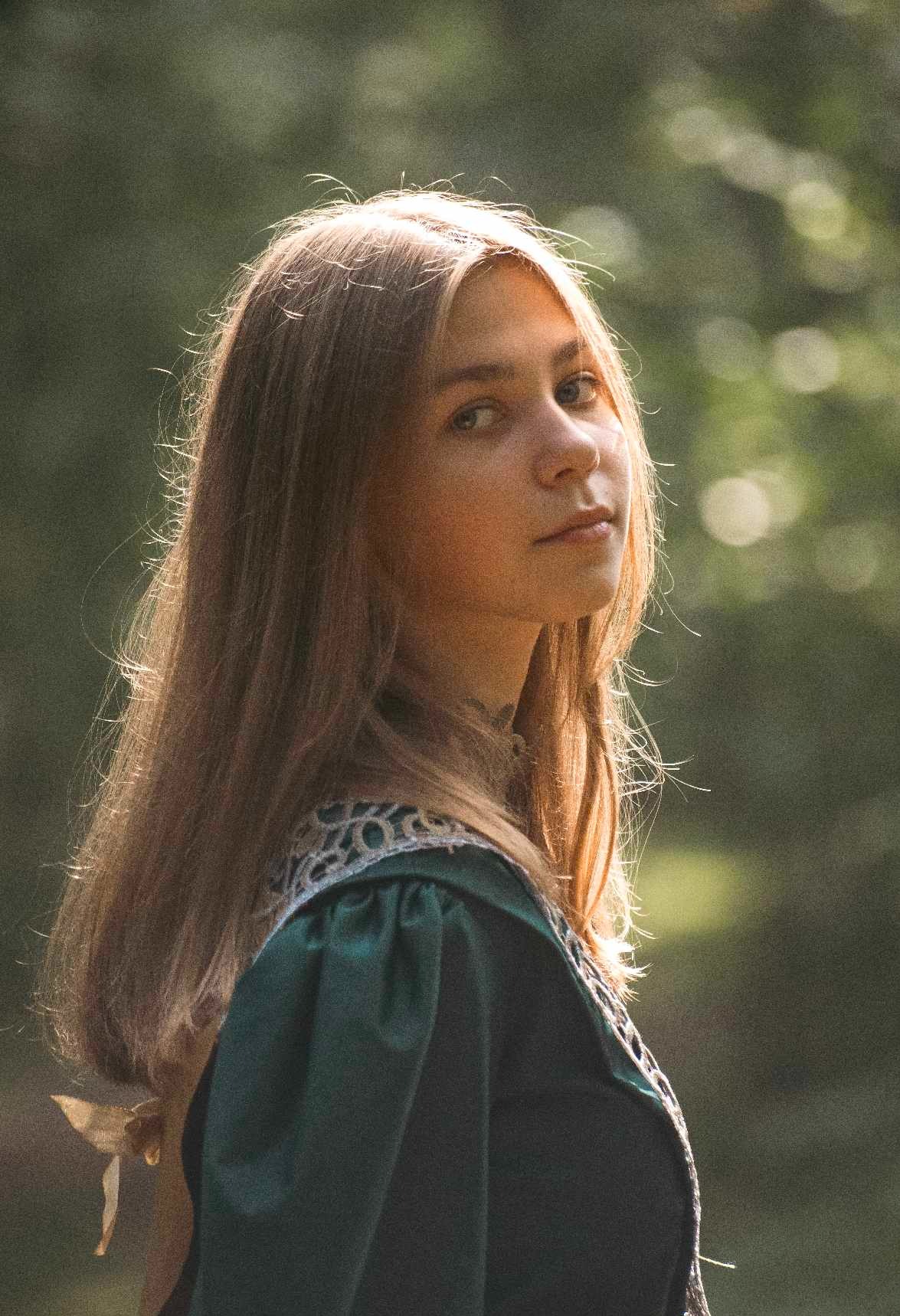 Portret młodej kobiety z długimi włosami, odwróconej w zielonej sukni z koronkowym wykończeniem, z delikatnym oświetleniem i rozmytym tłem.