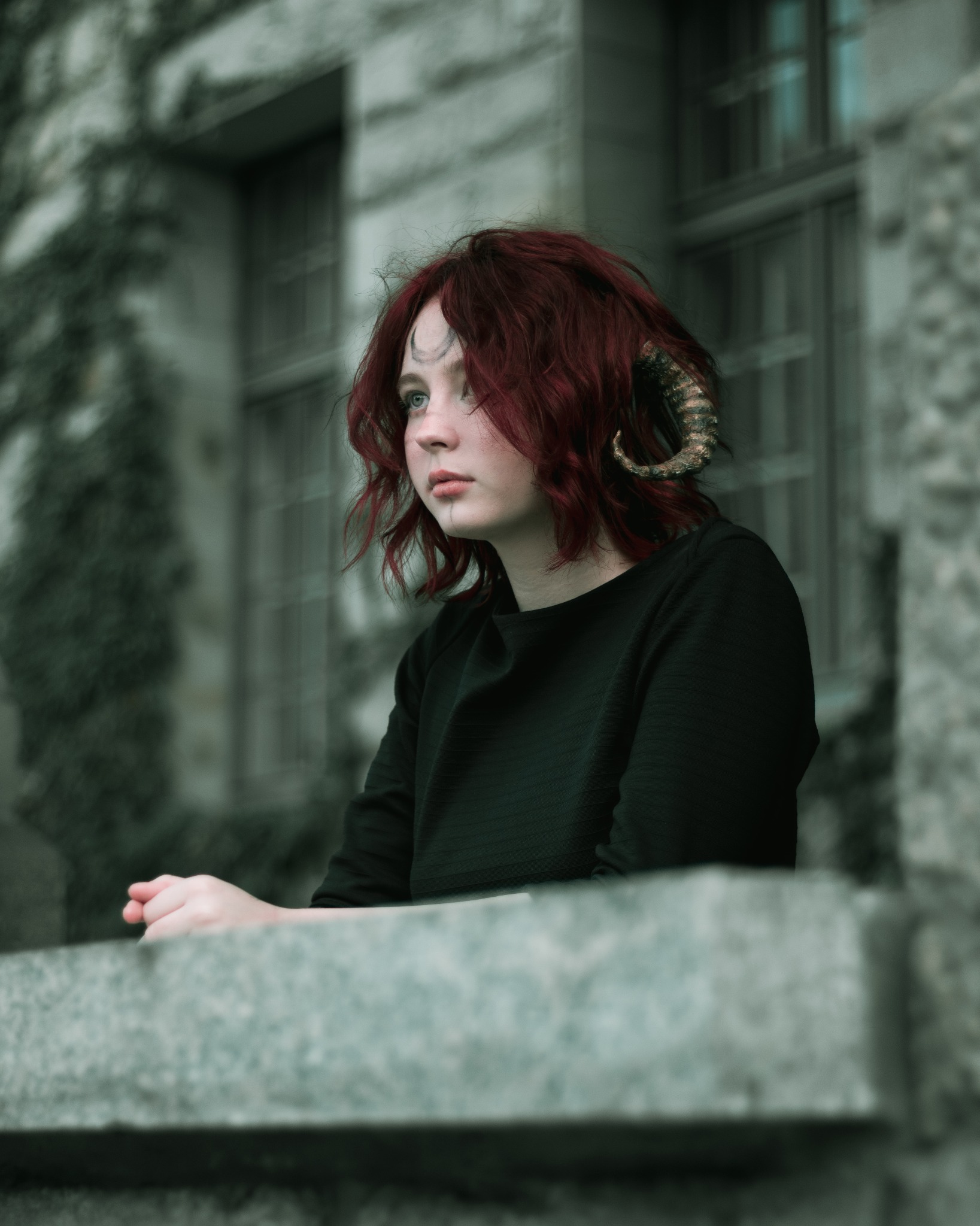 Portret kobiety z rudymi włosami, delikatnym makijażem, tatuażem na czole i ozdobnymi rogami, stojącej na tle kamiennej ściany z oknami.