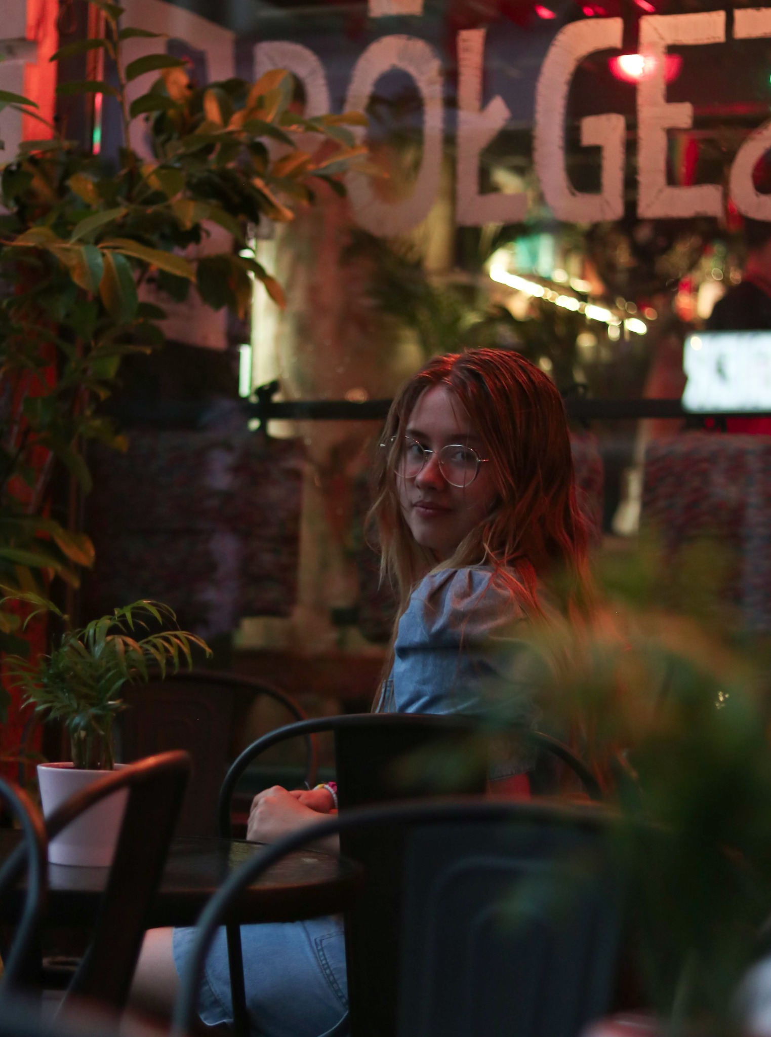 Portret młodej kobiety w okularach, siedzącej przy stoliku w kawiarni, z widocznymi roślinami i neonowym napisem za nią, wykonany w ciepłym oświetleniu.
