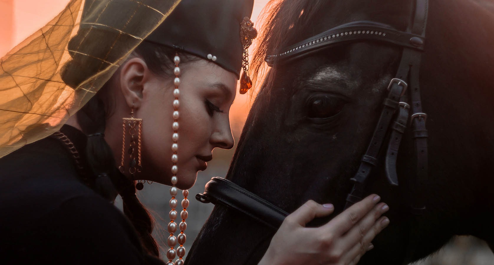 Kobieta w stylizowanej sukni i biżuterii, delikatnie dotyka pyska czarnego konia, ciepłe światło zachodzącego słońca.