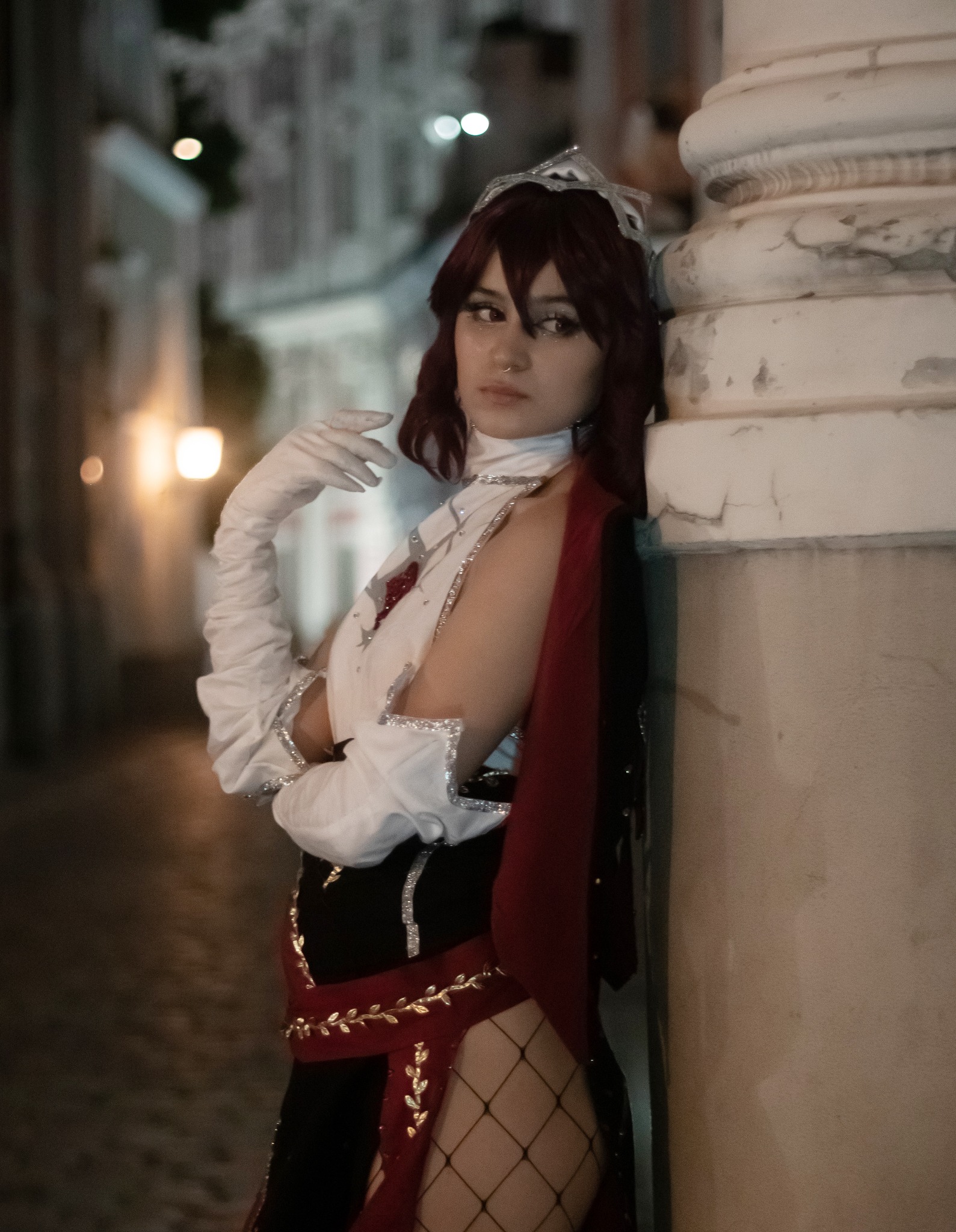 Kobieta w kostiumie królowej, oparta o kamienną kolumnę na tle nocnej ulicy, z koroną, długimi białymi rękawiczkami i strojem z czerwonymi akcentami i złotymi zdobieniami.