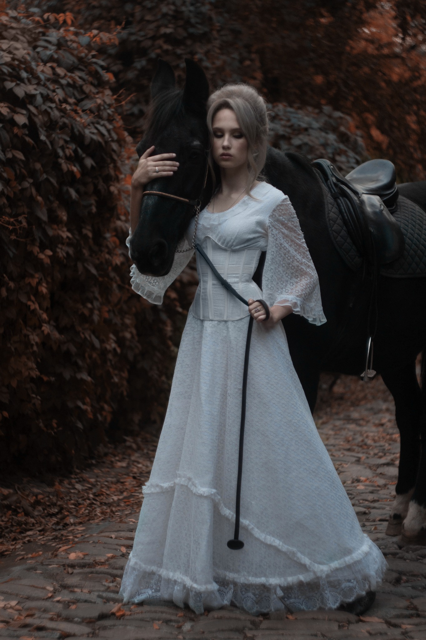 Kobieta w koronkowej sukni, z upiętymi blond włosami, obejmuje czarnego konia w jesiennej scenerii.