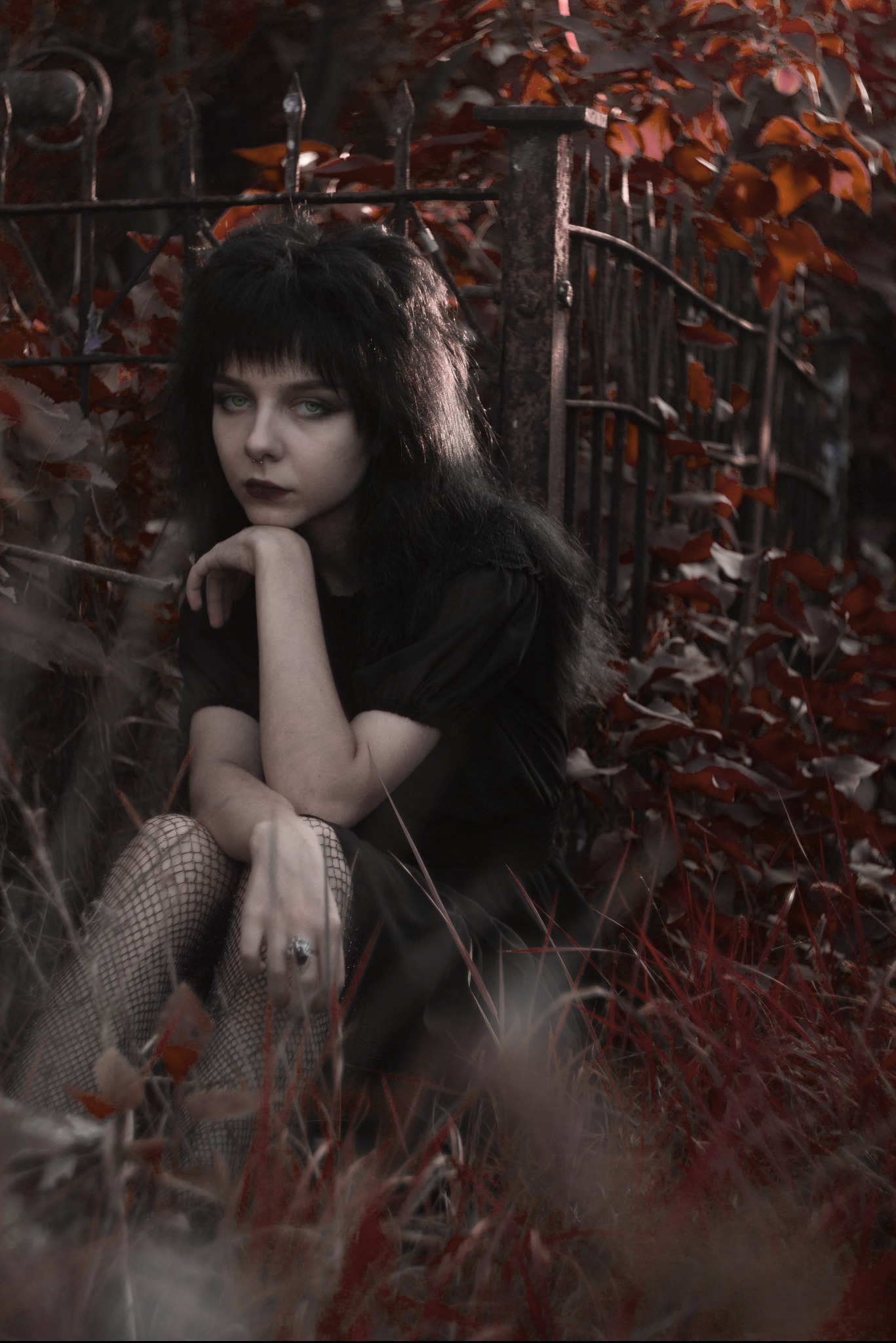 Portret kobiety w czarnej sukience i rajstopach w siatkę, siedzącej w trawie przed metalowym ogrodzeniem z jesiennymi liśćmi w tle, z wyrazistym makijażem i pierścionkiem na palcu.