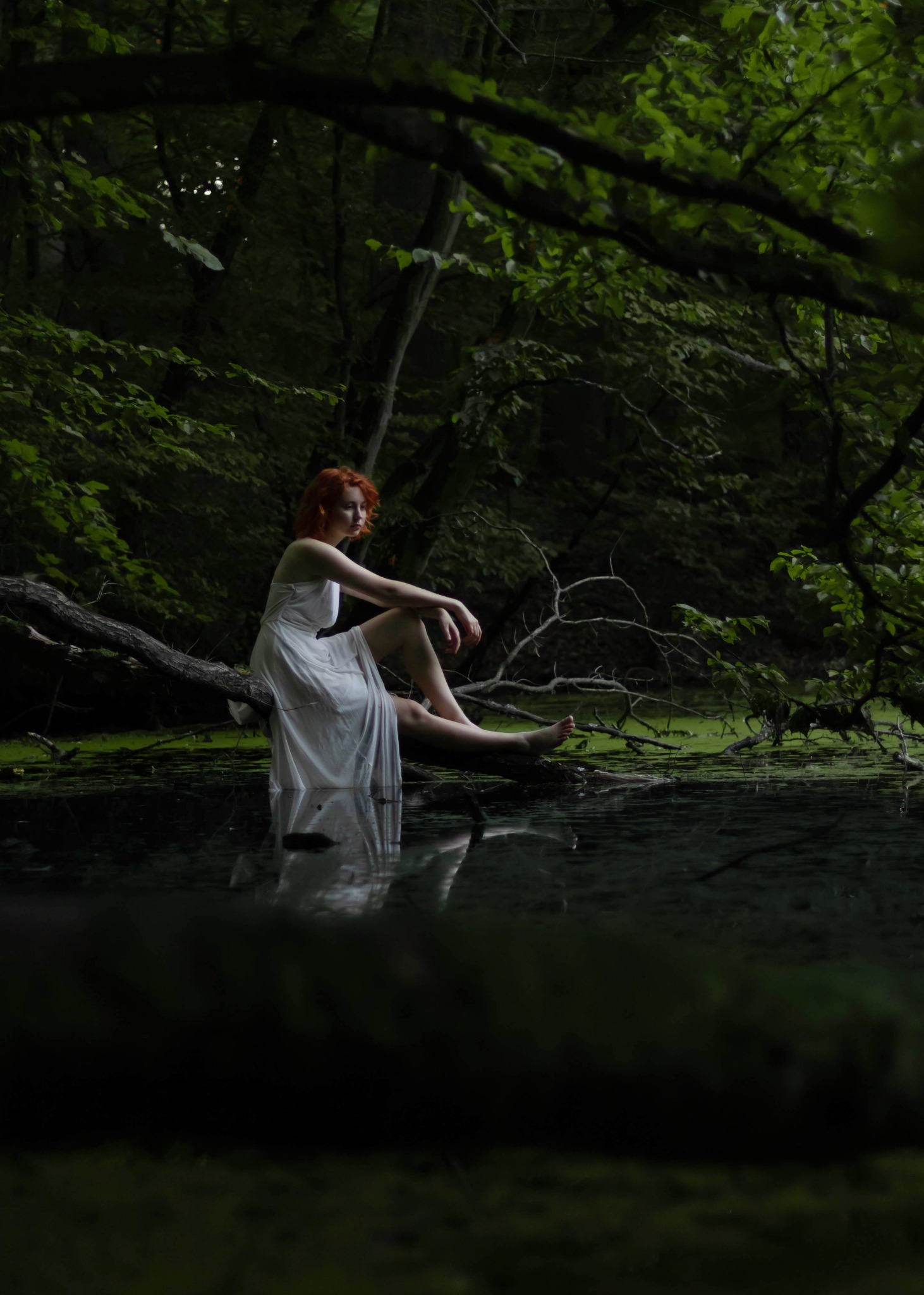 Rudowłosa kobieta w białej sukience siedzi boso na powalonym pniu drzewa nad wodą porośniętą rzęsą w zalesionym otoczeniu.